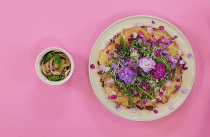 A hwajeon pizza topped with pink and purple flowers and greens sits on a white plate against a pink background, next to a side dish of marinated onions and peppers.
