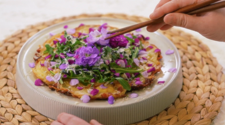 A hwajeon pizza topped with pink and purple flowers and greens sits on a white plate on top of a Korean straw mat, as a diner raises chopsticks to the flowers.