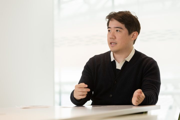 Image of a Asian man in a black sweater with white collar inside a white room and desk while in the middle of explaining something