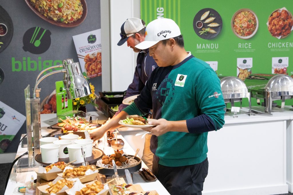 Two golfers holding a plate at a bibigo stand with foods in front