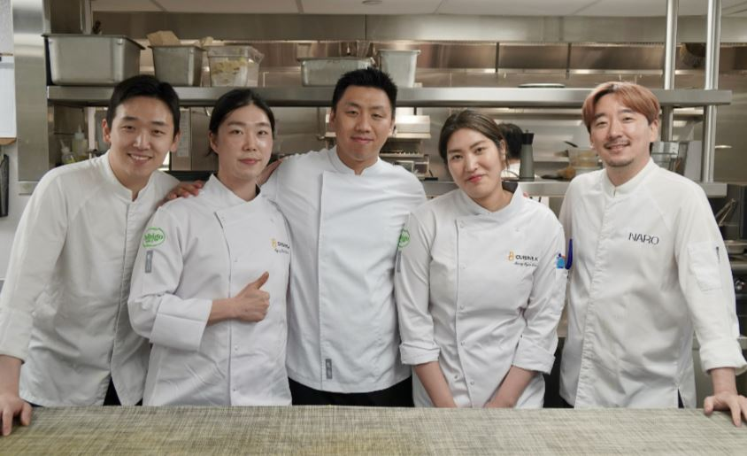 Chefs in white uniforms, three men and two women, smile for the camera in NARO’s kitchen.