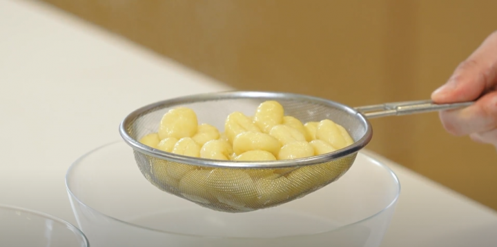 Strainer filled with gnocchi and a large clear bowl below. 
