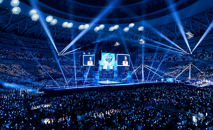 Wide view of a concert arena filled with thousands of fans and bright blue lights.
