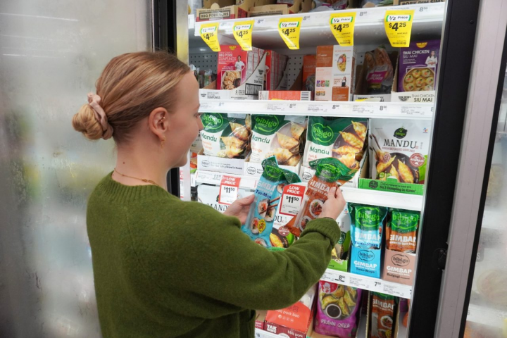 A woman in a green sweater compares two bibigo Gimbap packages in a grocery store freezer.
