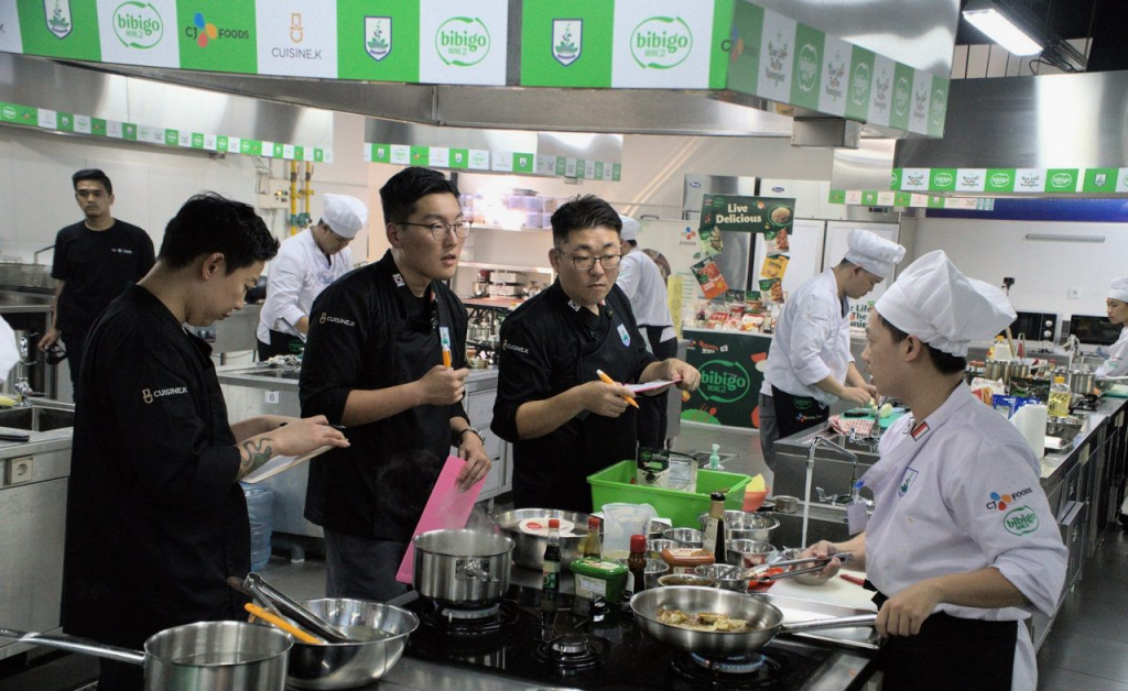 Three judges wearing black chefs’ uniforms judge a competitor chef who is cooking, with bibigo, Sages and CJ logos in the background.