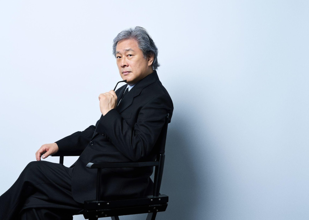 Director Park Chan-wook, dressed in a black coat, seated in a director’s chair.
