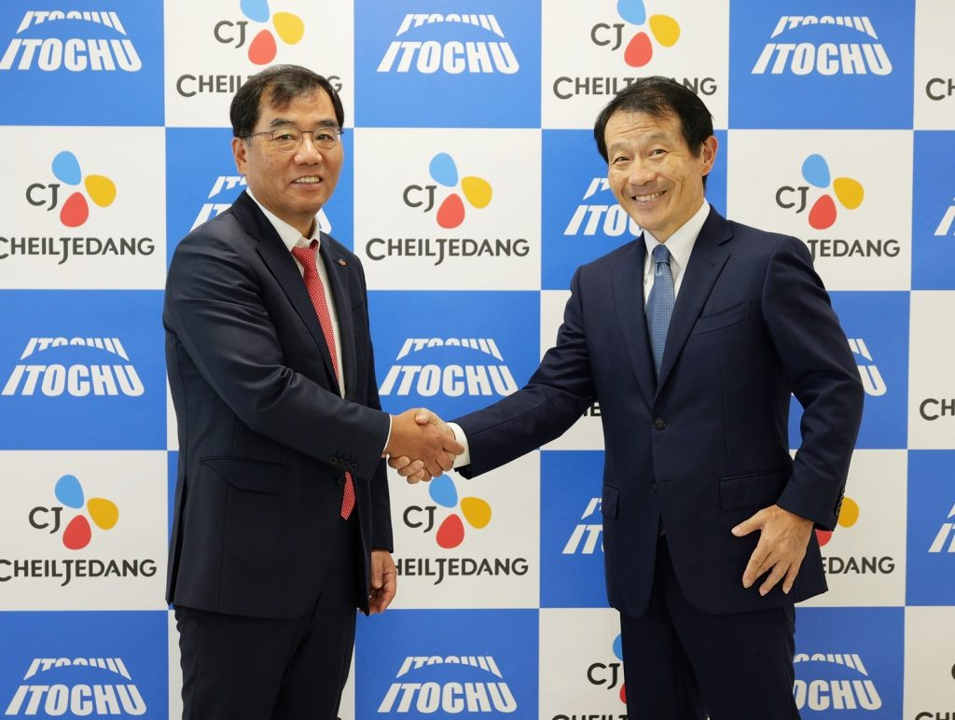 Two executives in business suits shaking hands in front of a CJ CheilJedang and ITOCHU logo wall. 
