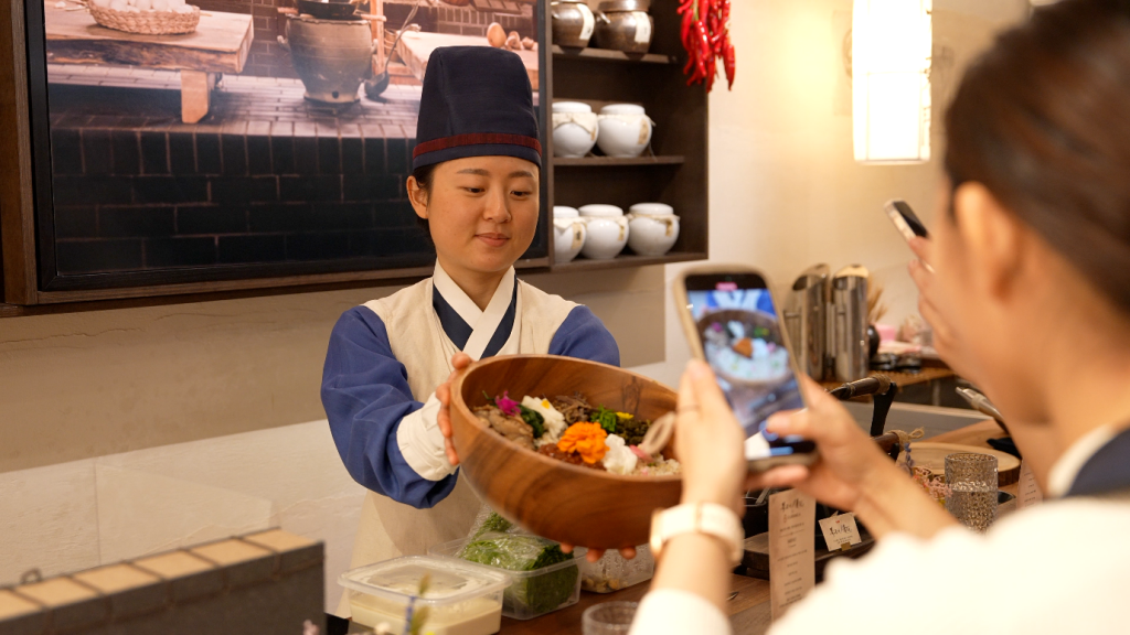 A chef dressed in Joseon garb holds a bibmbap dish as a diner takes a photo of it.