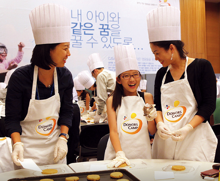 Two women and a young girl laugh as they bake cookies while wearing aprons and bakers’ hats at the 5th anniversary event for CJ Donorscamp.
