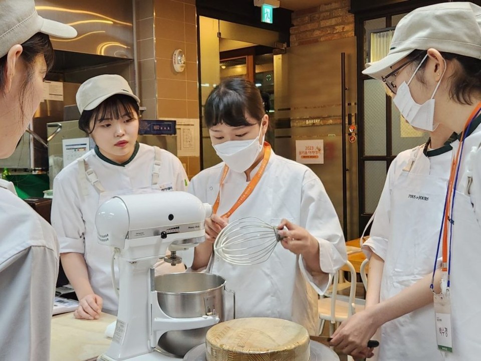 Four people gather around a baking stand while wearing baking uniforms for a CJ Donorscamp Academy training session.

