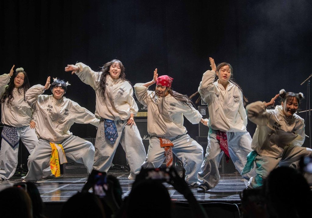 A group of six teenage girls dance on a stage wearing matching windbreakers and jeans.
