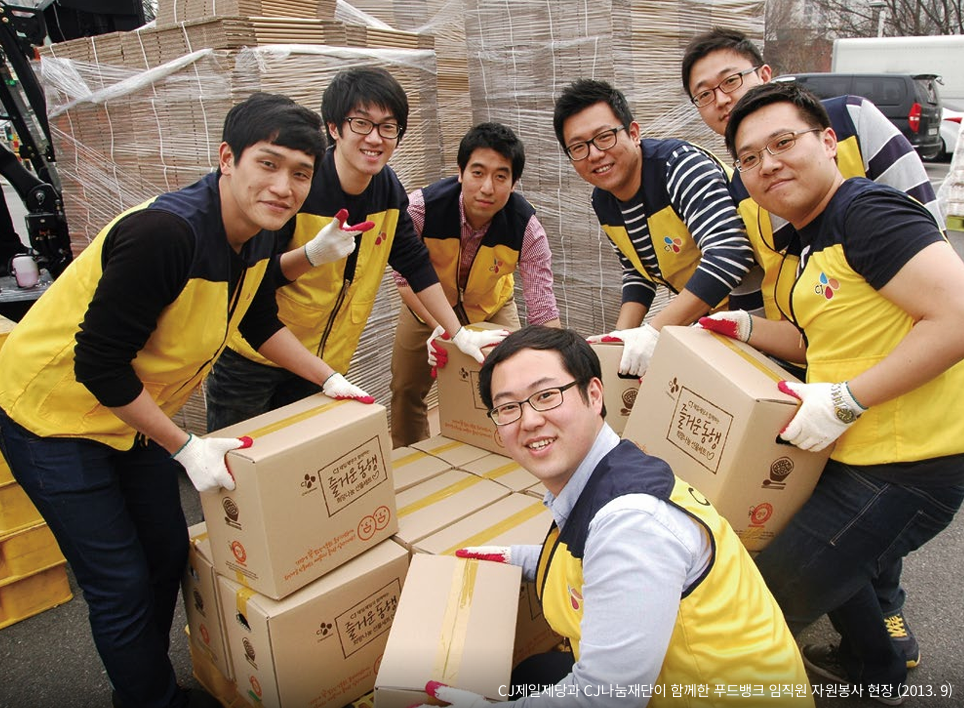 A group of seven men smile while wearing CJ uniform vests and holding cardboard boxes.
