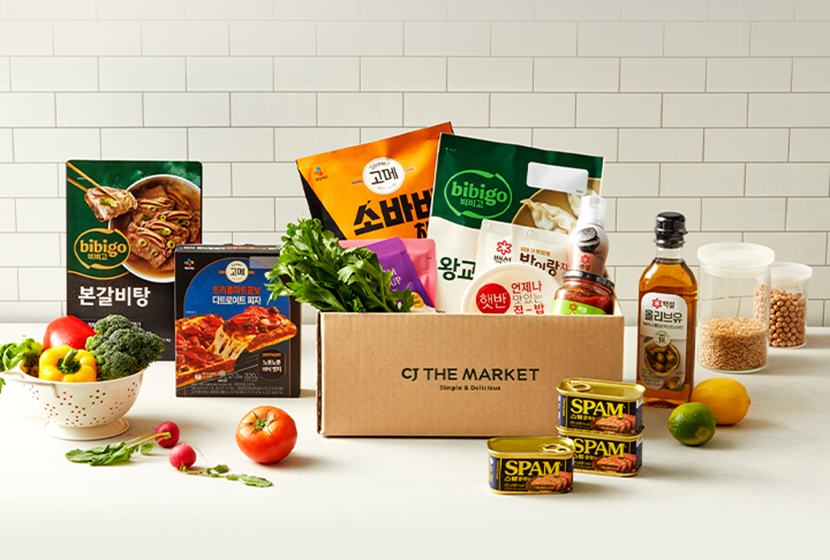 Several CJ Foods and bibigo products spread on a white countertop. Some products are in a CJ THE MARKET cardboard box.