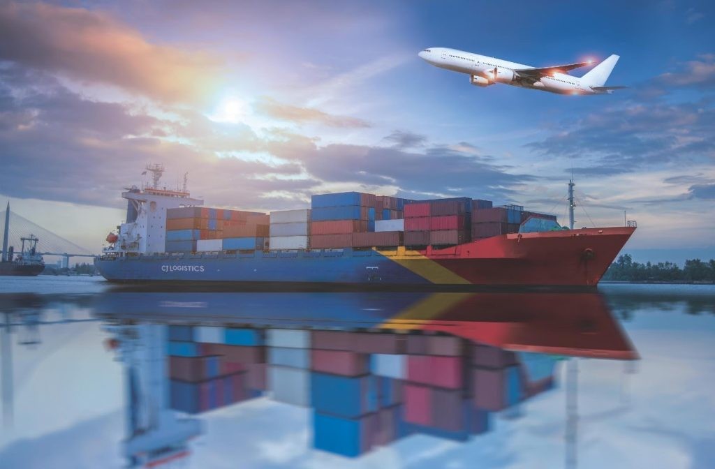 Image of a container ship with plane flying overheard the sun
