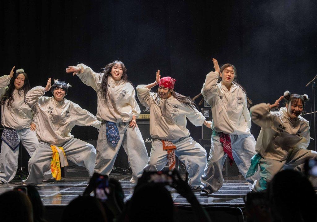 A group of six teenage girls dance on a stage wearing matching windbreakers and jeans.
