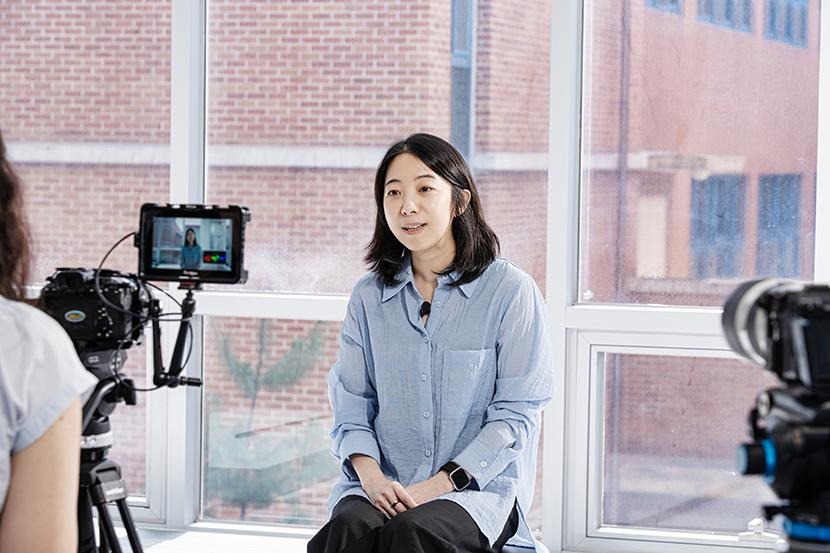 Against a window, Baek Hyun-jung sits and talks in front of a camera wearing a light-blue button-up shirt.
