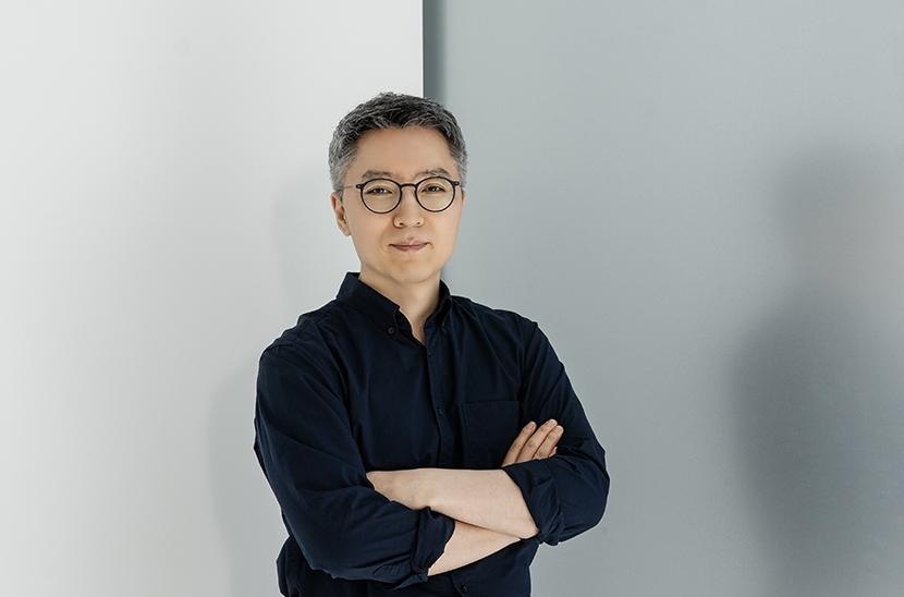 Against a white backdrop, Jeong Chang-ik crosses his arms while looking at the camera, wearing a black shirt with the sleeves rolled up.
