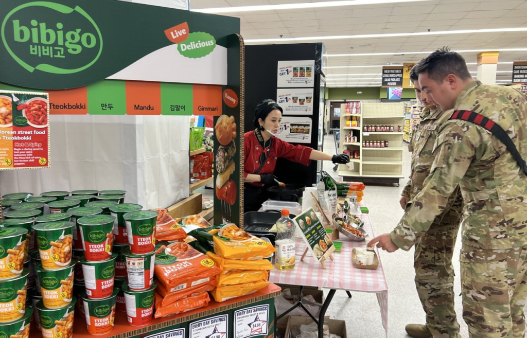 A CJ Foods rep offers bibigo tteokbokki samples to U.S. service members at a Guam military base commissary, highlighting K-food expansion.