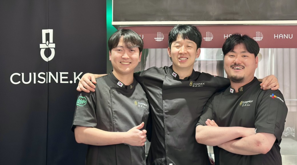 Three male chefs from Team Cuisine.K pose and smile in front of a Cuisine.K cloth
