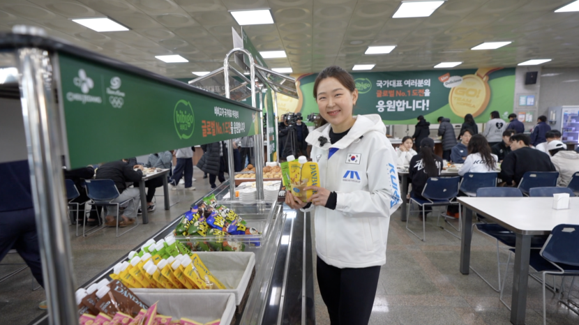 Kim Min-sun stands in a bibigo-sponsored cafeteria holding products and smiling.