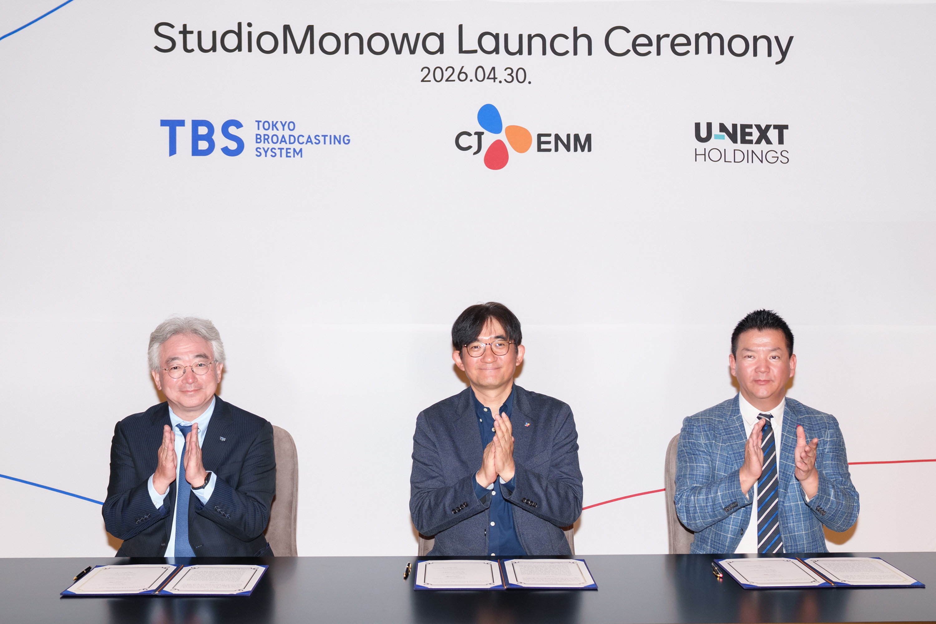 Three executives sit at a signing table during the StudioMonowa launch ceremony, with TBS, CJ ENM and U-NEXT Holdings logos displayed on the backdrop.