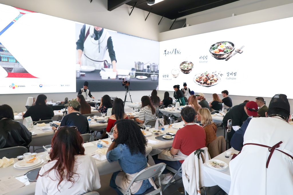 Participants seated at tables watch a Cuisine.K cooking demonstration on large screens.