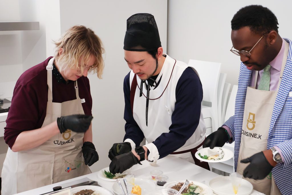 A chef guides two participants as they plate a dish during a cooking class.