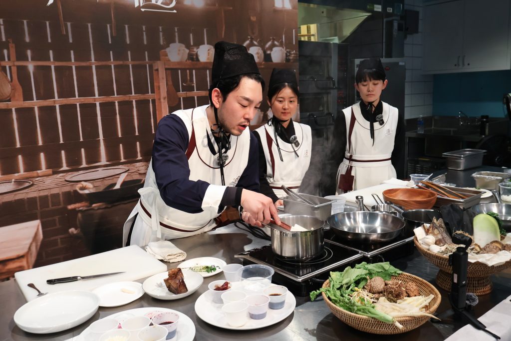 A Cuisine.K chef cooks at a station with ingredients laid out as two assistants watch.