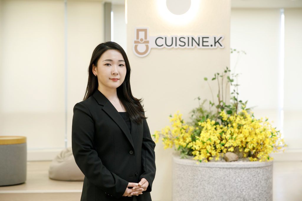 Park Shin-young poses in front of a Cuisine.K logo affixed to a wall
