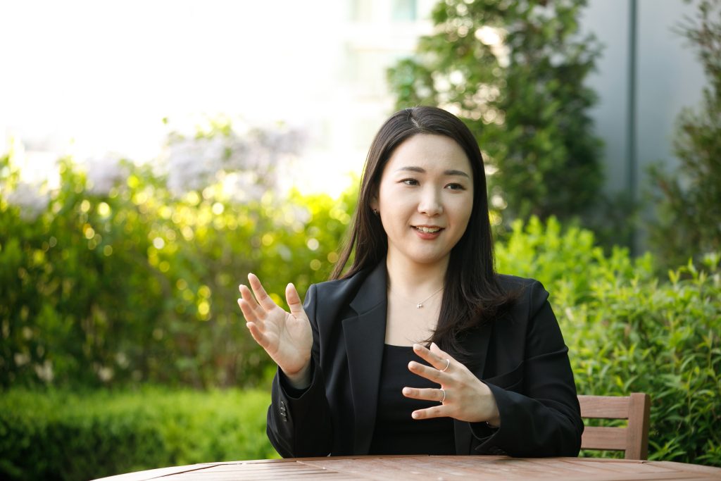 Park Shin-young sits at a table outside in front of shrubs