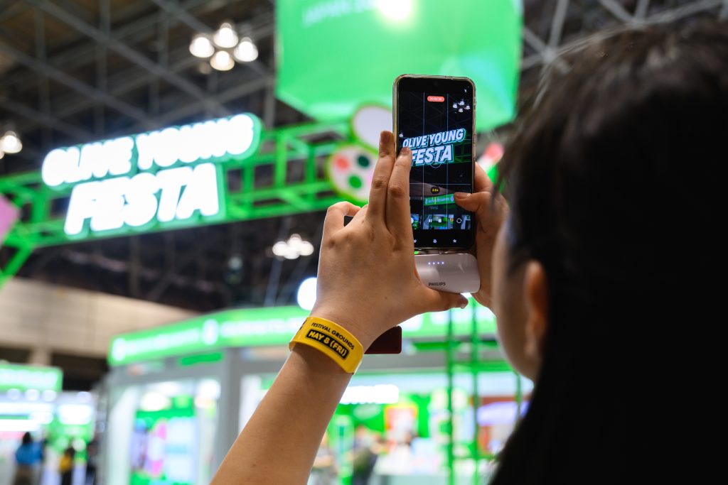 A visitor takes a photo of the OLIVE YOUNG FESTA JAPAN 2026 booth with a smartphone inside Makuhari Messe.