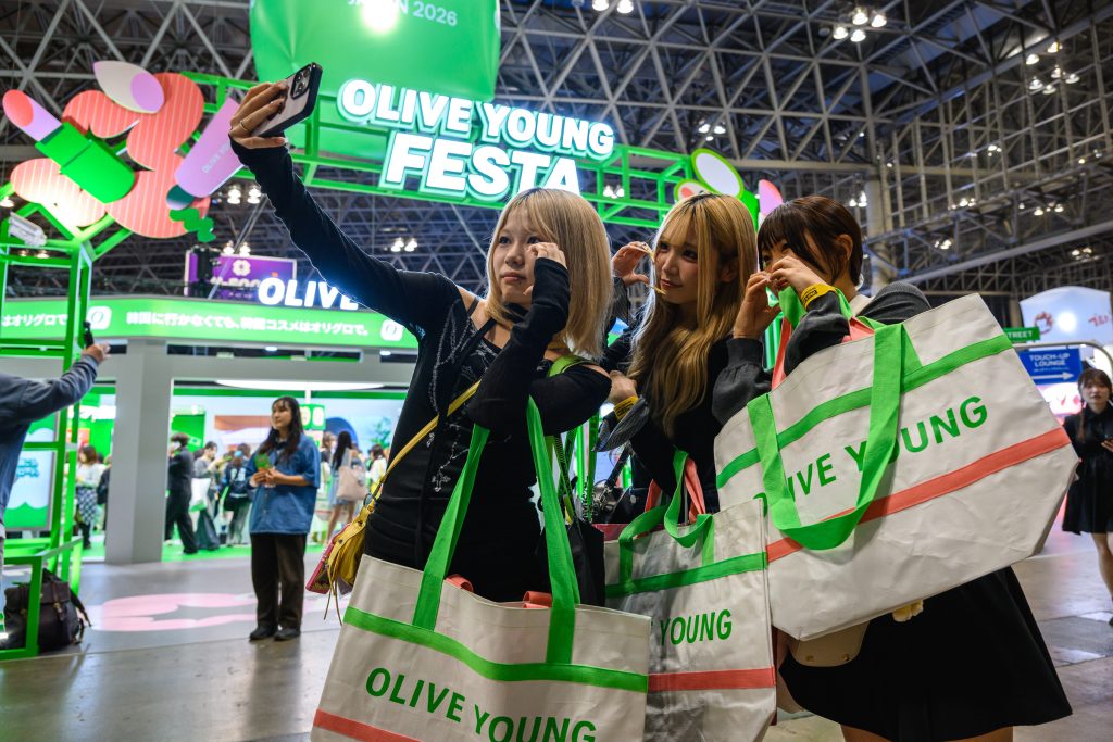 Three visitors take a selfie while holding OLIVE YOUNG shopping bags in front of the OLIVE YOUNG FESTA JAPAN 2026 booth.