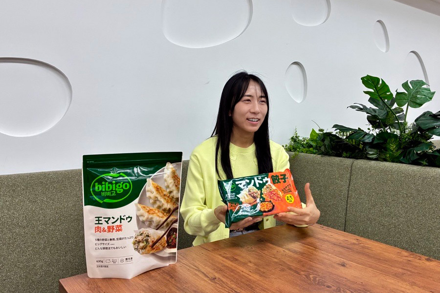 Ichikawa Sayo of CJ FOODS JAPAN speaks during an interview while holding bibigo Mandu Gyoza, with bibigo Korean-style mandu products displayed on the table.