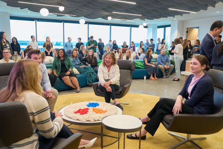 A large group of people sit throughout an open room on chairs and sofas and chat with one another.