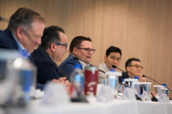 Five male executives sit on one side of a table, speaking into microphones.