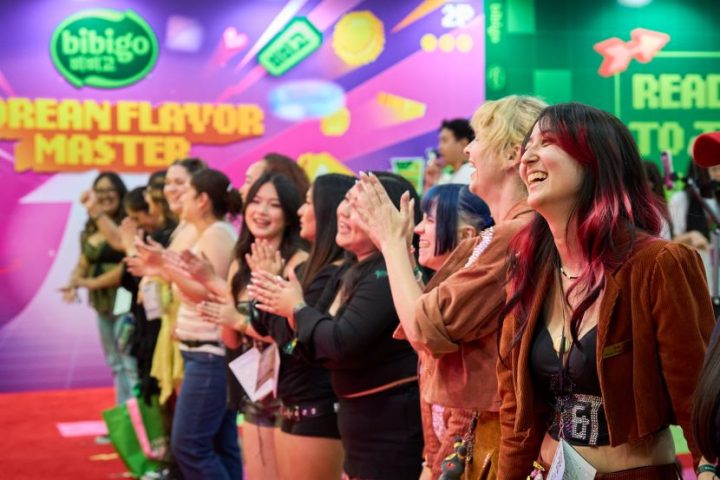 A group of people smile and clap, while in the background is a large bibigo advertisement with the title “KOREAN FLAVOR MASTER.”