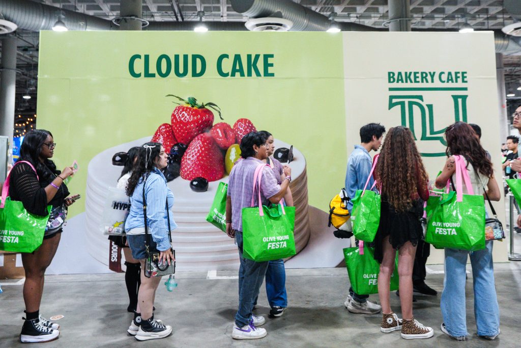 A group of people with OLIVE YOUNG FESTA shopping bags stand in front of a TOUS les JOURS advertisement for Cloud Cake.