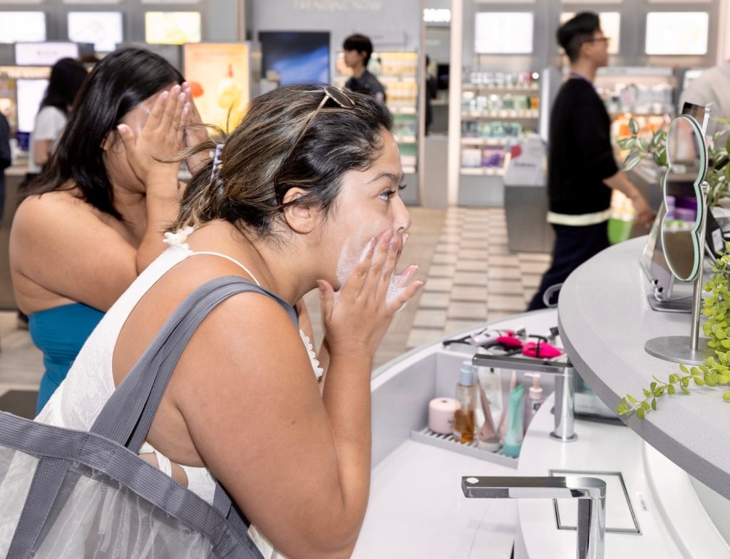 Two customers at OLIVE YOUNG wash their faces with a foam cleanser at a washing station.