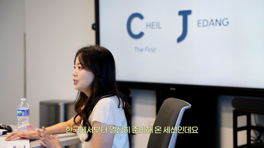 Lee speaks in front of a monitor in an office while sitting at a desk.